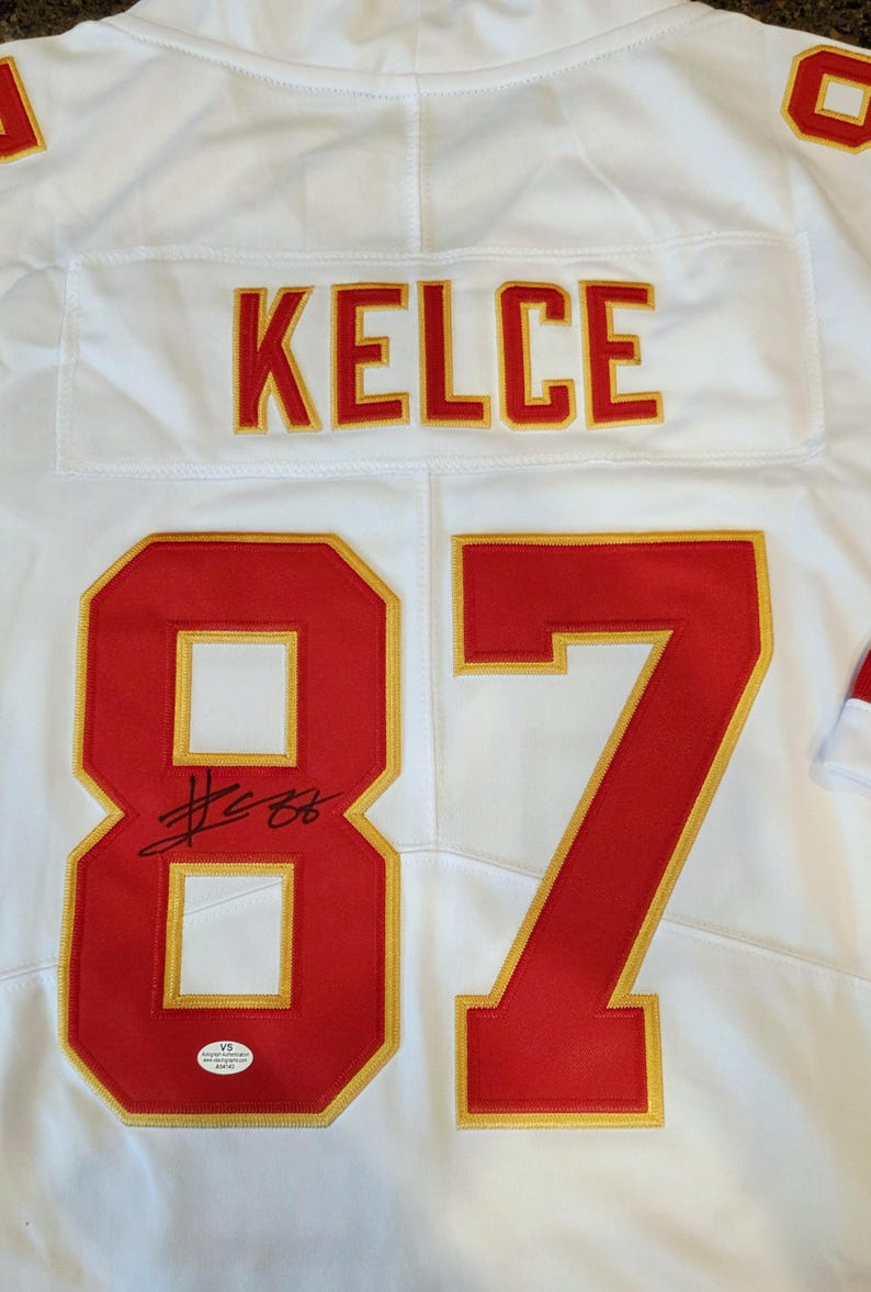 May include: White football jersey with the name "KELCE" in red and gold lettering. The number "87" is prominently displayed in red with gold trim. Includes an autograph and a certification sticker.