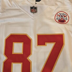 May include: White football jersey with red and gold accents. Features the number 87 in red with gold trim. Includes the NFL logo and a team logo patch. The jersey has a V-neck design.