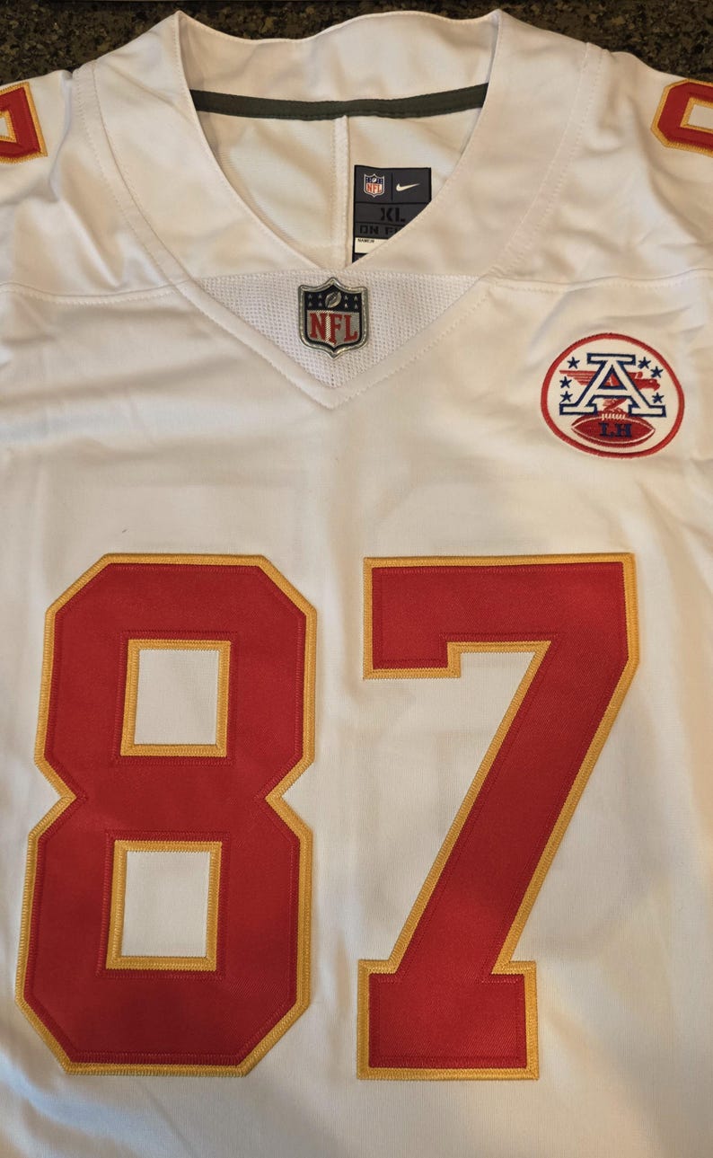 May include: White football jersey with red and gold accents. Features the number 87 in red with gold trim. Includes the NFL logo and a team logo patch. The jersey has a V-neck design.