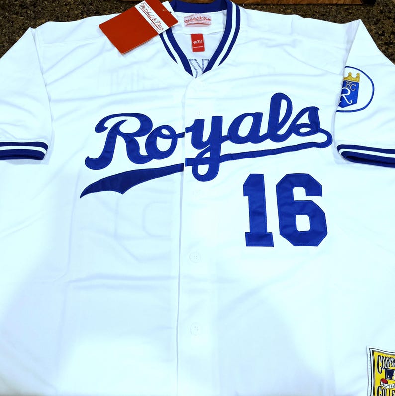 Puede incluir: Camiseta de b&eacute;isbol blanca con detalles azules, con la palabra "Royals" en cursiva y el n&uacute;mero "16". La camiseta tiene una parte delantera abotonada, un cuello en V y una etiqueta de Mitchell & Ness.