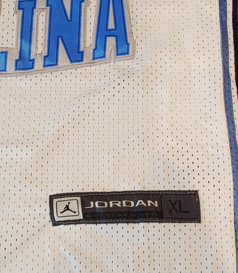 May include: White mesh jersey with blue accents and the word "INA" in blue. Features a "JORDAN" label with the size "XL". The jersey has a perforated design.