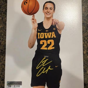 Op de afbeelding: Een glanzende foto van een basketbalspeler in een zwart Iowa Hawkeyes-shirt met het nummer 22 in het geel, die een oranje basketbal op één vinger laat draaien. De afbeelding bevat een gouden handtekening op de broek en een authenticatiesticker.