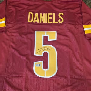 May include: A burgundy football jersey featuring the name DANIELS in yellow lettering above the number 5 in yellow with a white outline. The number 5 includes a black signature and a small silver authentication hologram sticker at the base.