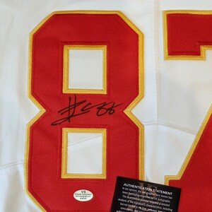 May include: White jersey with red and gold accents. Features the number 87 in red with a gold outline, and a black signature. Includes an authentication sticker and statement.