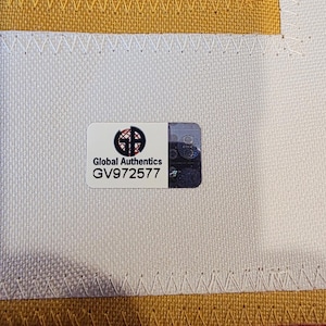 May include: Close-up of a jersey with a Global Authentics sticker. The jersey has a white base with red and gold accents. The sticker has the text "Global Authentics" and the serial number GV972577.