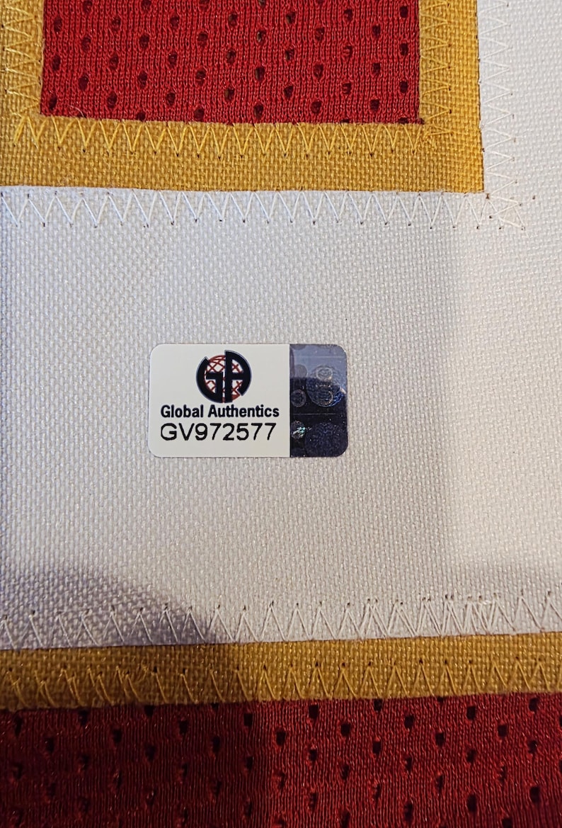 May include: Close-up of a jersey with a Global Authentics sticker. The jersey has a white base with red and gold accents. The sticker has the text "Global Authentics" and the serial number GV972577.
