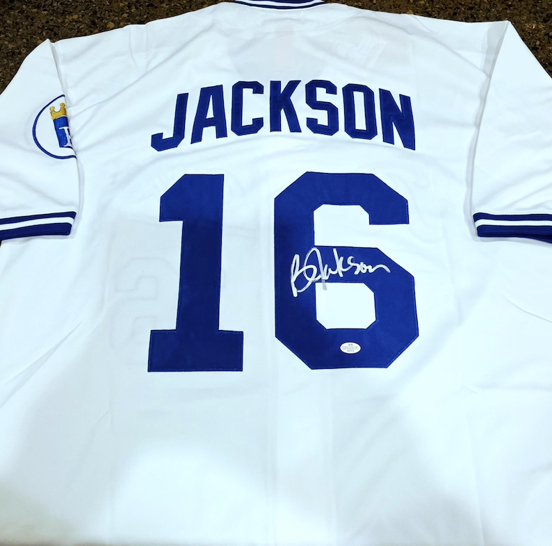 Puede incluir: Camiseta de b&eacute;isbol blanca con el nombre "JACKSON" en letras azules en la espalda y el n&uacute;mero "16" en azul. La camiseta tiene una firma y un logotipo del equipo en la manga.