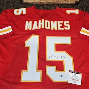 May include: A red football jersey with the name "MAHOMES" and the number "15" in white with gold trim. The jersey has a signature and a certification tag.