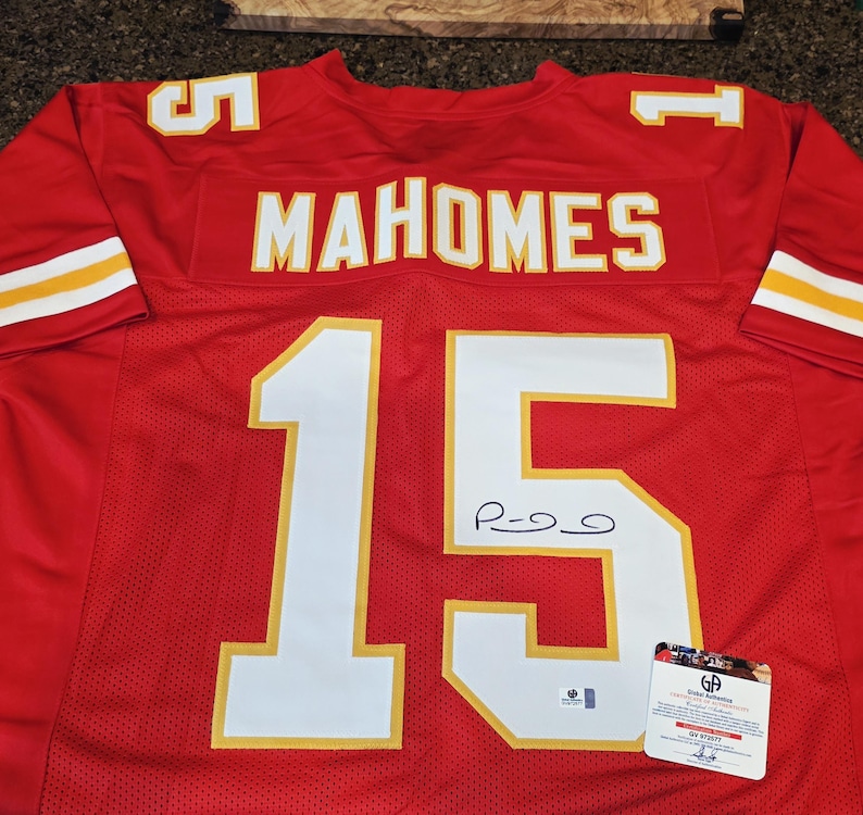 May include: A red football jersey with the name "MAHOMES" and the number "15" in white with gold trim. The jersey has a signature and a certification tag.