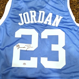 May include: Light blue basketball jersey with the name "JORDAN" and the number "23" in white. The jersey has a signature below the number. The jersey is made of a breathable mesh material.