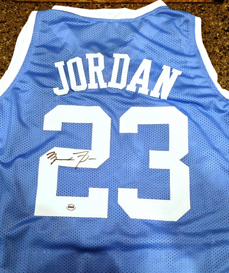 May include: Light blue basketball jersey with the name "JORDAN" and the number "23" in white. The jersey has a signature below the number. The jersey is made of a breathable mesh material.