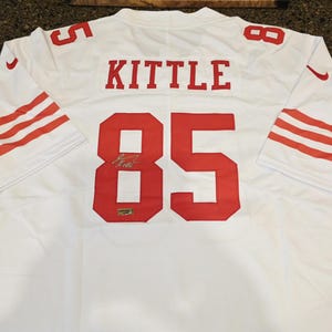 May include: A white sports jersey featuring the name KITTLE and the number 85 in red on the back. The sleeves have three red stripes, and a small red signature is visible on the number 8. A small rectangular authentication tag is on the number 8.