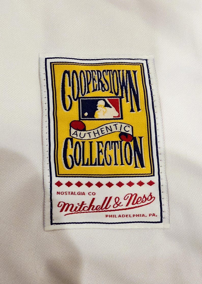 Puede incluir: Una etiqueta rectangular con las palabras "COOPERSTOWN AUTHENTIC COLLECTION" en letras azul marino y rojo. La etiqueta es amarilla con un logotipo de b&eacute;isbol rojo y azul. La marca "Mitchell & Ness" est&aacute; en escritura roja.
