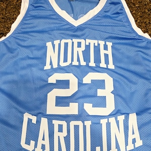 May include: Light blue basketball jersey with white trim and the words "NORTH CAROLINA" and the number "23" in white block letters. The jersey has a V-neck and is made of a breathable mesh fabric.