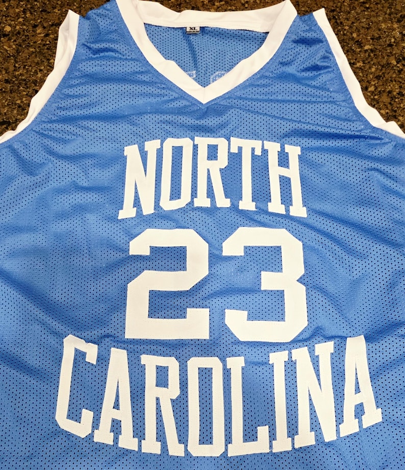 May include: Light blue basketball jersey with white trim and the words "NORTH CAROLINA" and the number "23" in white block letters. The jersey has a V-neck and is made of a breathable mesh fabric.