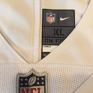 May include: Close-up of an NFL jersey with a white and olive green design. The jersey features the NFL logo, a Nike logo, and a size tag that reads XL. The jersey is made of a breathable mesh material.