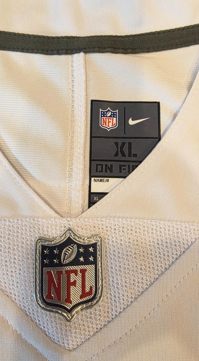 May include: Close-up of an NFL jersey with a white and olive green design. The jersey features the NFL logo, a Nike logo, and a size tag that reads XL. The jersey is made of a breathable mesh material.
