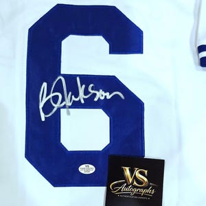 Puede incluir: Camiseta blanca con un gran n&uacute;mero 6 azul y una firma. La camiseta tambi&eacute;n tiene el nombre "BROOKS" en letras azules. En la imagen tambi&eacute;n hay una tarjeta negra con letras doradas que dice "VS Autographs".