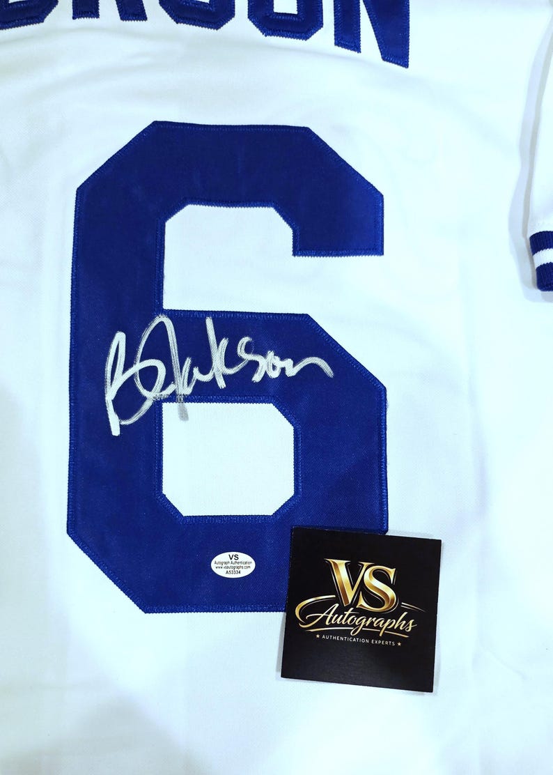 Puede incluir: Camiseta blanca con un gran n&uacute;mero 6 azul y una firma. La camiseta tambi&eacute;n tiene el nombre "BROOKS" en letras azules. En la imagen tambi&eacute;n hay una tarjeta negra con letras doradas que dice "VS Autographs".