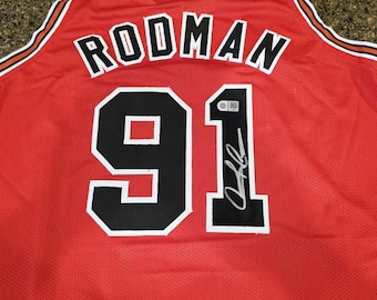 May include: A red basketball jersey featuring the name RODMAN and the number 91 in black with white outlines. The jersey has a mesh texture, striped trim at the neckline, and a black signature autograph on the number 91 with a small authentication tag.