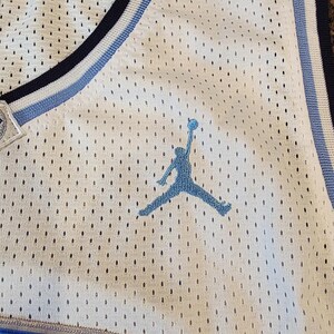 May include: White mesh basketball jersey with blue and light blue accents. Features a blue silhouette of a person jumping with a basketball. The jersey has a blue and white striped trim around the neckline and armholes.