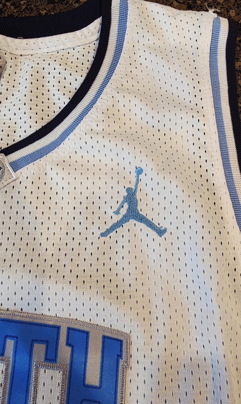 May include: White mesh basketball jersey with blue and light blue accents. Features a blue silhouette of a person jumping with a basketball. The jersey has a blue and white striped trim around the neckline and armholes.