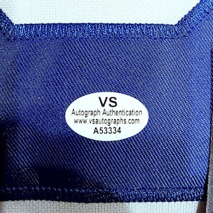 Puede incluir: Primer plano de una tela azul con una secci&oacute;n blanca en la parte superior. Una pegatina ovalada con el texto "VS Autograph Authentication www.vsautographs.com A53334" est&aacute; adherida a la tela azul.
