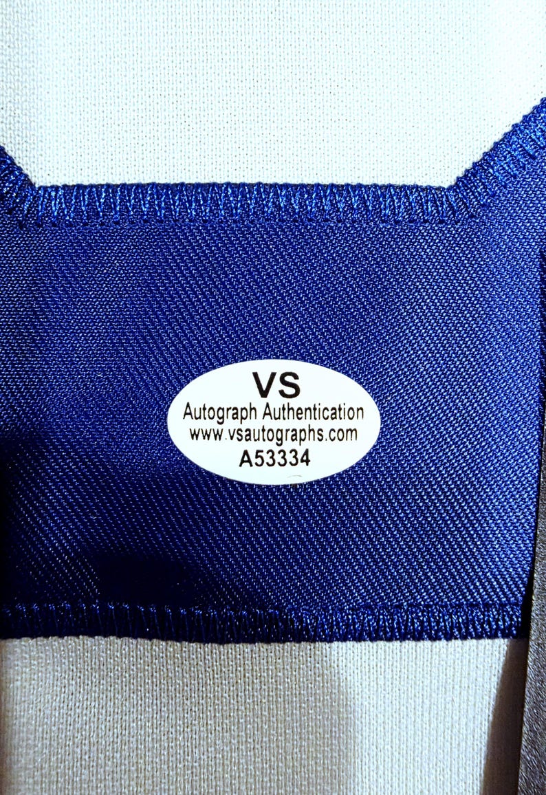 Puede incluir: Primer plano de una tela azul con una secci&oacute;n blanca en la parte superior. Una pegatina ovalada con el texto "VS Autograph Authentication www.vsautographs.com A53334" est&aacute; adherida a la tela azul.