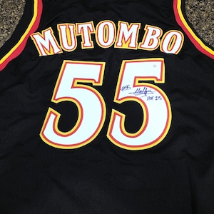 Può includere: Maglia da basket nera con il nome "MUTOMBO" sulle spalle in lettere bianche con bordi gialli. Il numero "55" è visualizzato in grandi numeri bianchi con bordi gialli. La maglia presenta una firma e la scritta "HOF 15".