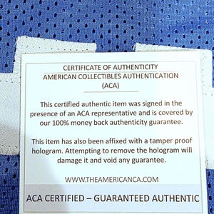 May include: A certificate of authenticity from American Collectibles Authentication (ACA) is displayed. The white card states the item is certified authentic, signed in the presence of an ACA representative, and covered by a money-back guarantee. The card also mentions a tamper-proof hologram.