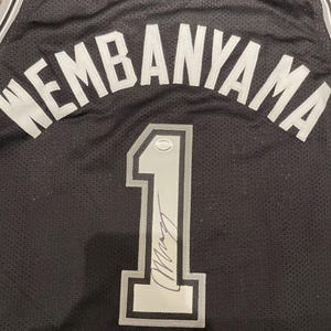 May include: Black basketball jersey with the name "WEMBANYAMA" in white lettering across the top. The number "1" is prominently displayed in silver with a signature below. The jersey has a mesh texture.