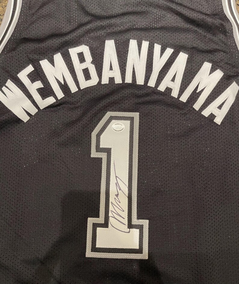 May include: Black basketball jersey with the name "WEMBANYAMA" in white lettering across the top. The number "1" is prominently displayed in silver with a signature below. The jersey has a mesh texture.
