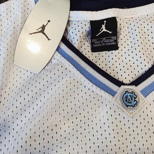 May include: White mesh jersey with a V-neck design, featuring light blue and navy blue accents. The jersey has a tag with the Jordan logo and a patch with the letters "UNC". The jersey is made in Thailand.
