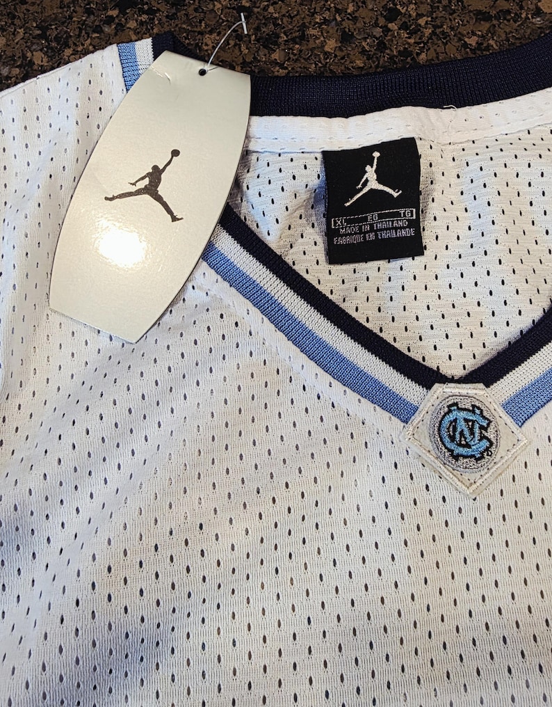 May include: White mesh jersey with a V-neck design, featuring light blue and navy blue accents. The jersey has a tag with the Jordan logo and a patch with the letters "UNC". The jersey is made in Thailand.