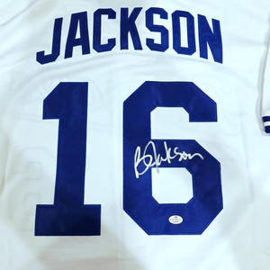 Puede incluir: Camiseta de b&eacute;isbol blanca con el nombre "JACKSON" en letras azules en la espalda. El n&uacute;mero "16" tambi&eacute;n es azul, con una firma debajo. La camiseta tiene ribetes a rayas azules y blancas.