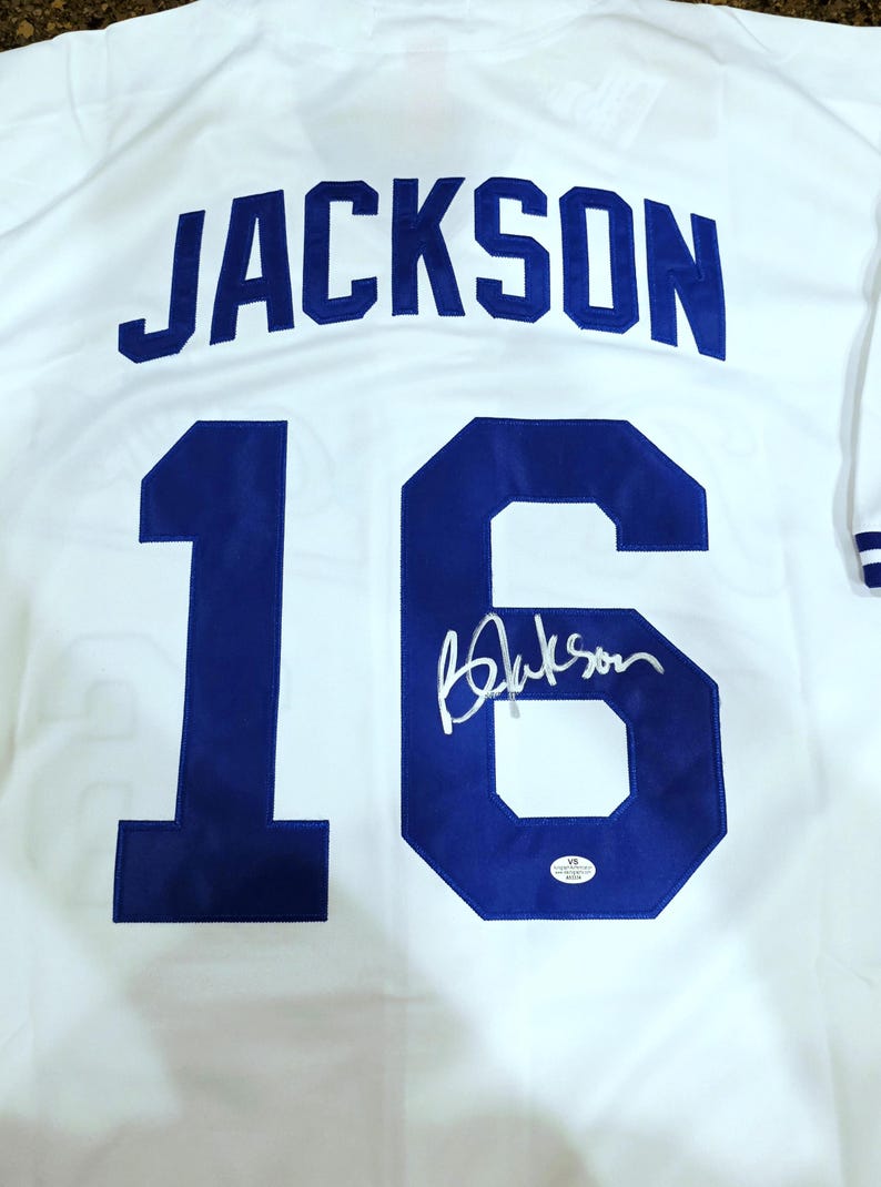 Puede incluir: Camiseta de b&eacute;isbol blanca con el nombre "JACKSON" en letras azules en la espalda. El n&uacute;mero "16" tambi&eacute;n es azul, con una firma debajo. La camiseta tiene ribetes a rayas azules y blancas.