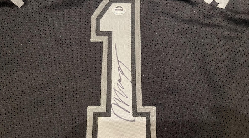 May include: Black jersey with a large, white and gray number 1 on the front. The number is outlined in gray and has a signature across it. An American Authentication sticker is visible.