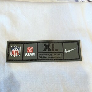 May include: A rectangular patch with the NFL logo, NFL Players logo, and the text "XL" and "ENGINEERED TO THE EXACT SPECIFICATIONS OF THE NATIONAL FOOTBALL LEAGUE" and a Nike swoosh. The patch is on a white fabric.