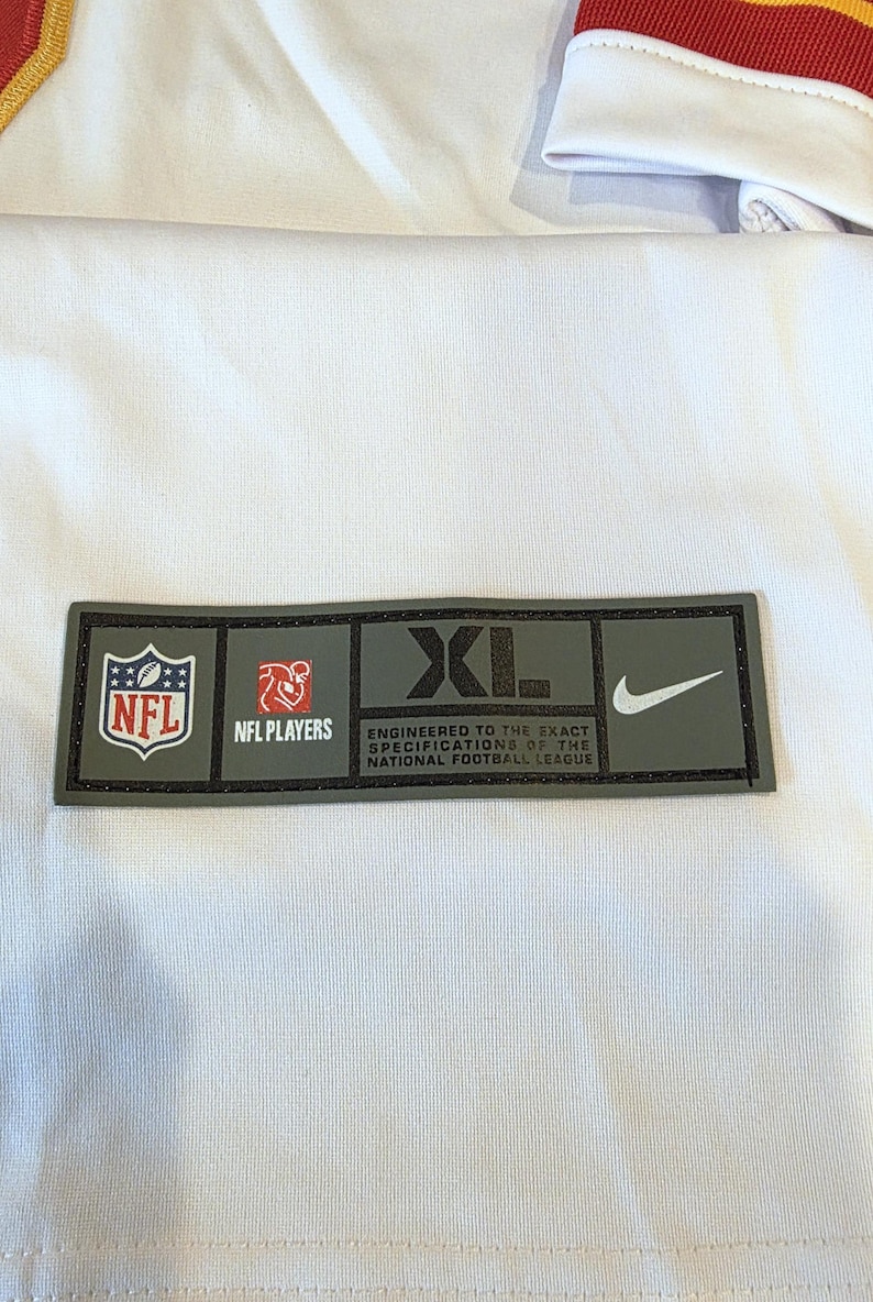 May include: A rectangular patch with the NFL logo, NFL Players logo, and the text "XL" and "ENGINEERED TO THE EXACT SPECIFICATIONS OF THE NATIONAL FOOTBALL LEAGUE" and a Nike swoosh. The patch is on a white fabric.