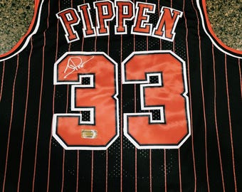 May include: A black basketball jersey featuring thin vertical orange pinstripes, orange trim on the collar and armholes, and the name PIPPEN with the number 33 in orange and white on the back. A signature is visible on the number 33.