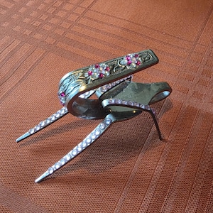May include: An ornate silver-toned capo with pink and clear rhinestones. The capo features a floral design with red accents and is designed to fit over the guitar strings. The capo is resting on a brown surface.