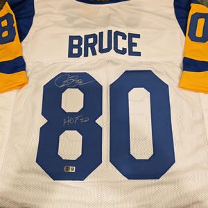 Puede incluir: Una camiseta de malla blanca con detalles azules y amarillos. La camiseta presenta el nombre "BRUCE" en el pecho y el número "80" en azul en la parte delantera y trasera. La camiseta está firmada.