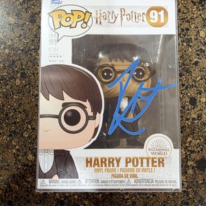 May include: A Funko Pop! vinyl figure of Harry Potter, number 91, in its original packaging. The box features a cartoon image of Harry Potter with glasses and a signature in blue ink. The packaging includes the text "Harry Potter" and "Wizarding World."