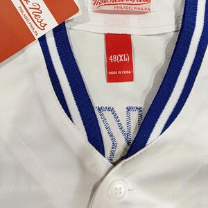 Puede incluir: Camiseta de b&eacute;isbol blanca con ribete a rayas azules y blancas en el cuello y las mangas. La camiseta tiene una parte delantera abotonada y una etiqueta roja que dice "48 (XL)" y "Made in China". El logo de Mitchell & Ness tambi&eacute;n est&aacute; presente.
