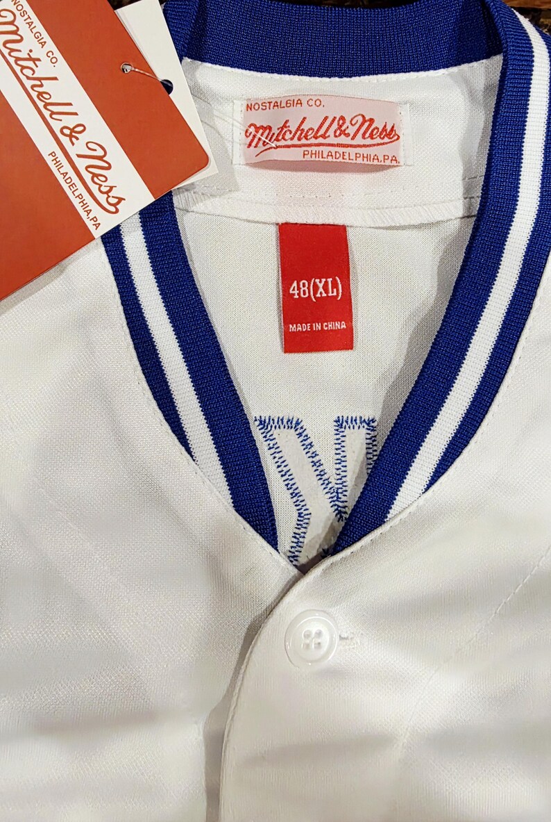 Puede incluir: Camiseta de b&eacute;isbol blanca con ribete a rayas azules y blancas en el cuello y las mangas. La camiseta tiene una parte delantera abotonada y una etiqueta roja que dice "48 (XL)" y "Made in China". El logo de Mitchell & Ness tambi&eacute;n est&aacute; presente.