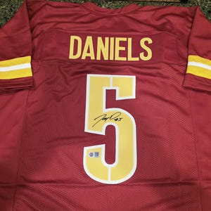 May include: A burgundy sports jersey featuring the name DANIELS in yellow lettering across the upper back. A large yellow number 5 is centred below the name, featuring a black ink signature and a small rectangular authentication sticker at the base.
