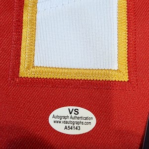 May include: Close-up of a red fabric with a white and gold embroidered patch. The patch is rectangular with a white center and gold trim. A white oval sticker with black text reads "VS Autograph Authentication www.vsautographs.com A54143".