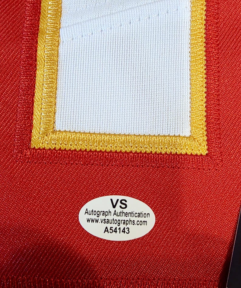 May include: Close-up of a red fabric with a white and gold embroidered patch. The patch is rectangular with a white center and gold trim. A white oval sticker with black text reads "VS Autograph Authentication www.vsautographs.com A54143".
