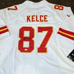 May include: White football jersey with red and gold accents. Features the name "KELCE" and the number "87" in red. Includes a signature and a Nike tag.