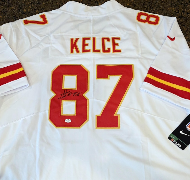 May include: White football jersey with red and gold accents. Features the name "KELCE" and the number "87" in red. Includes a signature and a Nike tag.
